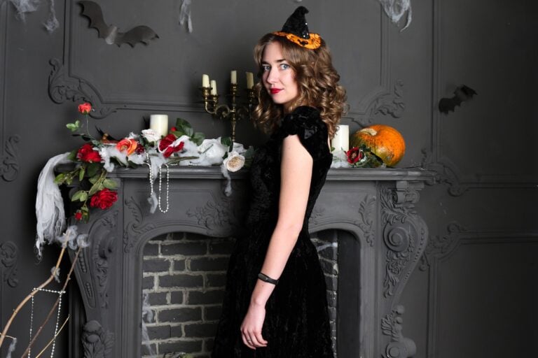 Woman wearing a black dress and witch hat beside a Halloween-decorated fireplace